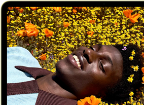MacBook Neo, citrus colour, screen shows a smiling person lying in a sunny field of flowers