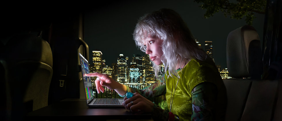 A person sitting in the back seat of a van, the open passenger door reveals a city skyline at night, MacBook Pro with PROLED XDR Display on a worktable, the person is illuminated by the screen while using the Touch feature to edit a video in DaVinci Resolve Studio