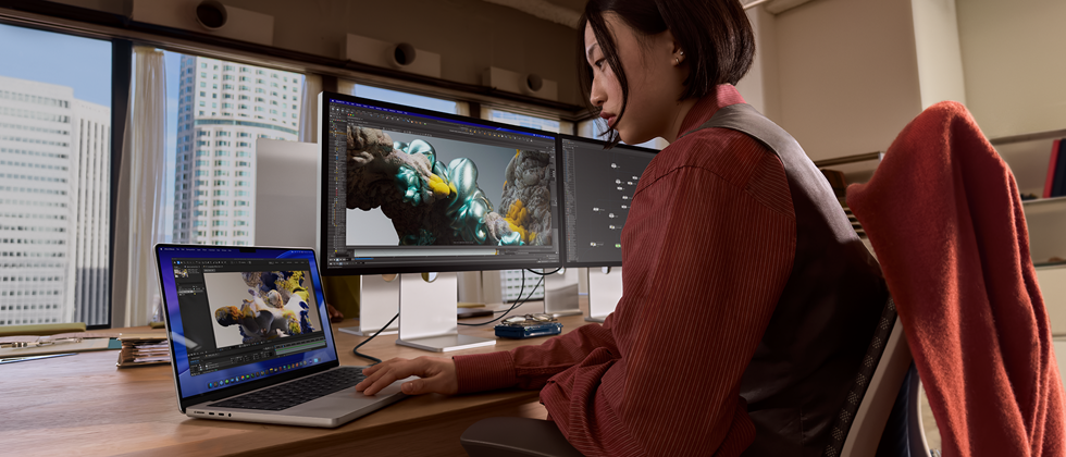 A person working in an office setting with tall buildings visible through windows, with MacBook Pro connected to Studio Display XDR