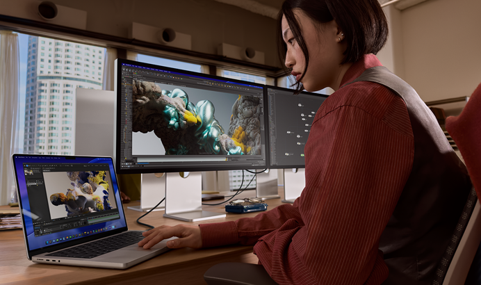 A person working in an office setting with tall buildings visible through windows, with MacBook Pro connected to Studio Display XDR