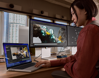 A person working in an office setting with tall buildings visible through windows, with MacBook Pro connected to Studio Display XDR